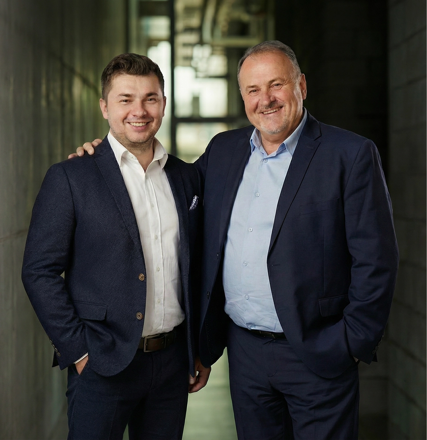 Dwóch uśmiechniętych mężczyzn w garniturach na tle nowoczesnego korytarza biurowego